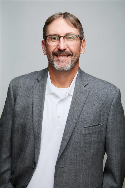 Bradley W. Smith, smiling, in a gray tweed blazer and white polo against a gray studio backdrop